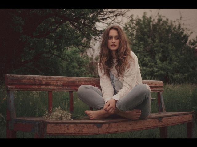 Alesya Drons sitzt barfuß auf einer rustikalen Holzbank, die Beine gekreuzt, und trägt eine weiße Spitzenbluse und graue Jeans. Sie hat langes, braunes Haar und befindet sich in einer grünen, offenen Umgebung..
