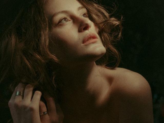 AlesyaDrons a les cheveux bruns ondulés, une épaule dénudée et une expression pensive avec les lèvres légèrement entrouvertes. Sa main, ornée de bagues, touche son cou. L'éclairage est tamisé et crée une ambiance mélancolique..