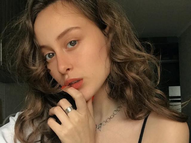 Alesya Drons mit welligem, braunem Haar, heller Haut und dezentem Make-up, die mit einem schwarzen Telefon in der Hand an ihrem Finger nagt, trägt ein schwarzes Top mit Trägern und einen silbernen Ring..