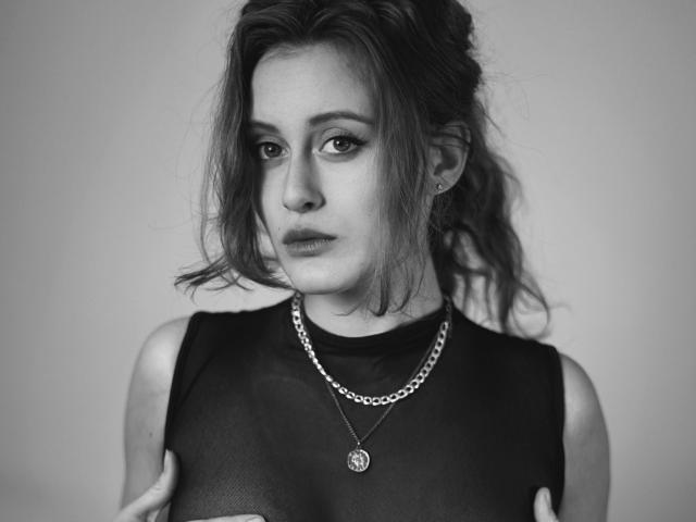 AlesyaDrons with messy hair, wearing a sheer black top, showing small tits, silver chain necklace, and a round pendant, looking down with a neutral expression, in black-and-white.