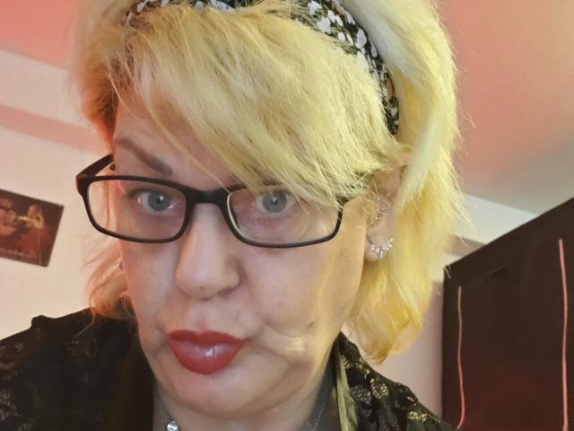 LatexShantie has blonde hair, black glasses, red lipstick, and a black-and-white headband, with a slightly pouty expression, wearing a dark, patterned top.