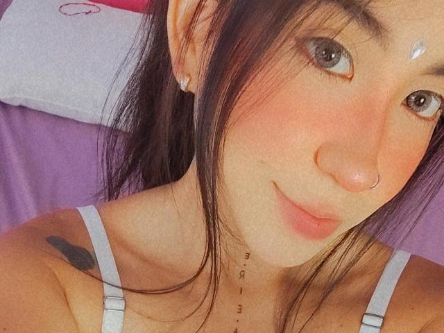Sarita Johnsons Gesicht mit langen, dunklen Haaren, einem Nasenring, hellrosa Lippen, grauen Augen und einem weißen Top mit Spaghetti-Trägern, wobei ein kleines, schwarzes Tattoo an ihrer rechten Schulter zu sehen ist..