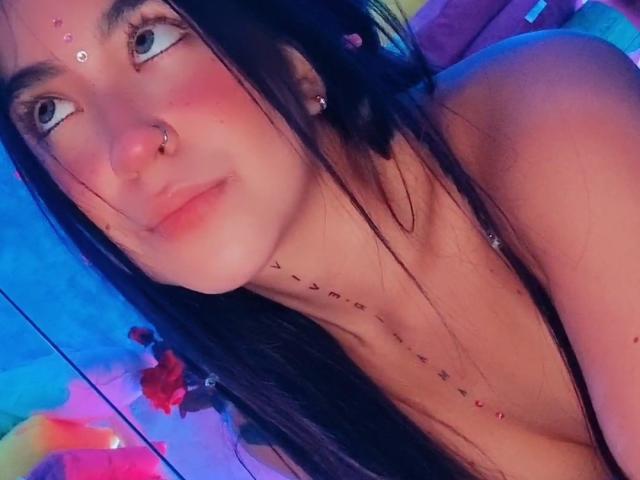 SaritaJohson with long black hair, nose ring, and facial jewelry, blue eyes, pink cheeks, necklace, and slight smile, showing bare shoulders. Her tits are partially visible.