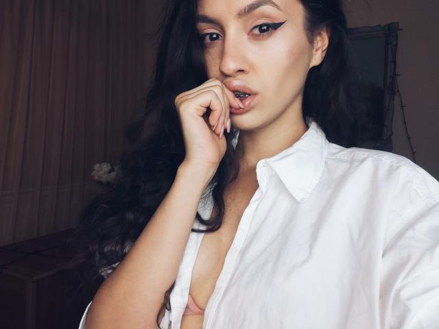 Vievienne Clemente, avec de longs cheveux noirs, chemise blanche ouverte, dévoilant sa poitrine, doigts près de la bouche, maquillage des yeux en forme d'ailes, regardant la caméra, dans un décor de chambre..