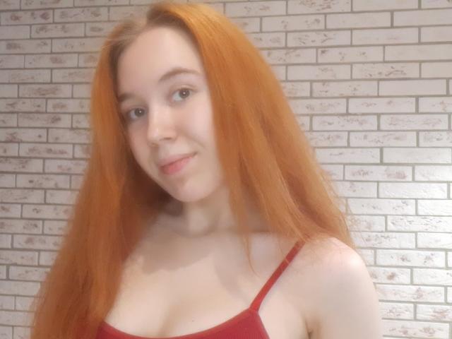 AnikaGlow has long red hair, light skin, and small tits. She's wearing a red bra, smiling slightly, with a soft, gentle expression. She looks cute and sexy.