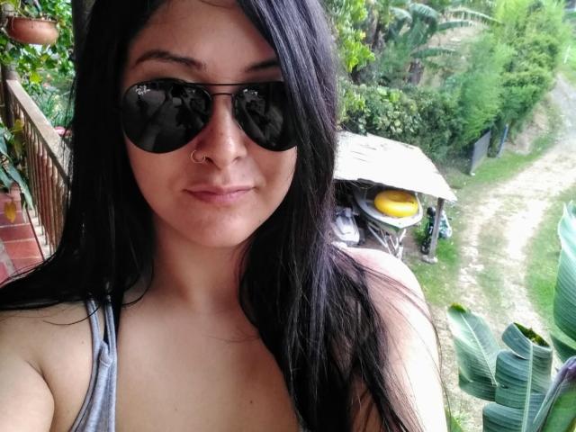 AnanovaX with long black hair, wearing dark sunglasses and a gray tank top, showing her nose ring and small tits, standing outdoors with a leafy background.