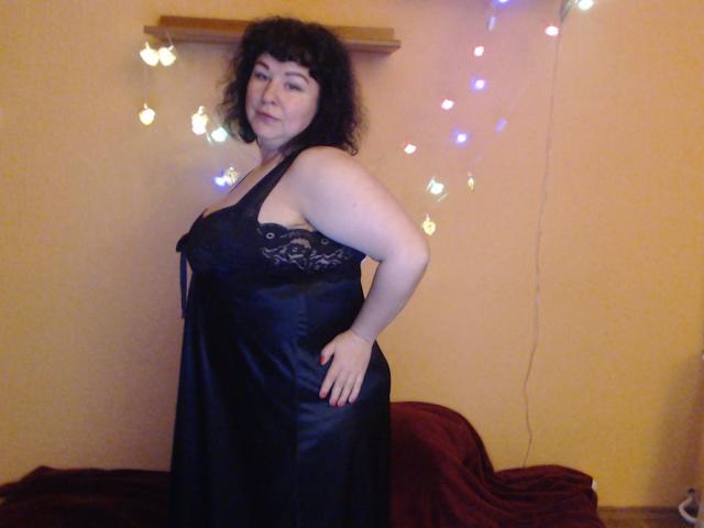 ErinSmart, avec ses cheveux noirs bouclés, portant une robe noire en dentelle et en satin, debout, une main sur sa hanche, montrant sa poitrine et ses courbes..