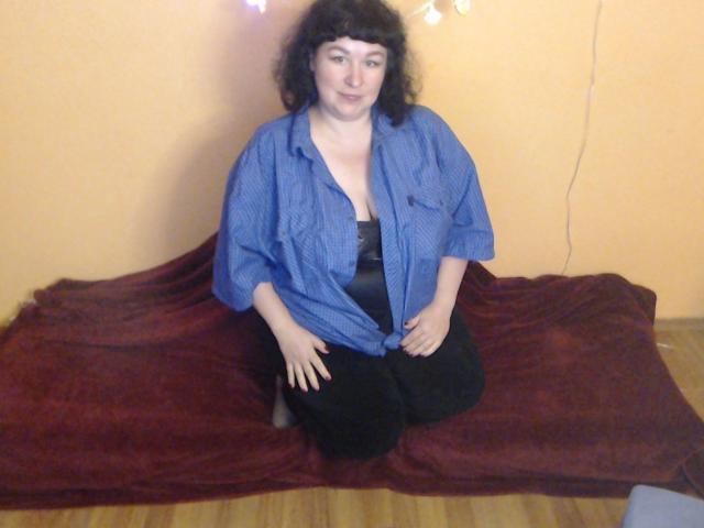 ErinSmart, avec ses cheveux noirs bouclés, portant une chemise bleue à boutons et un pantalon noir, est agenouillée sur un tissu rouge, montrant son soutien-gorge noir et ses petites seins. Le sol est en bois..