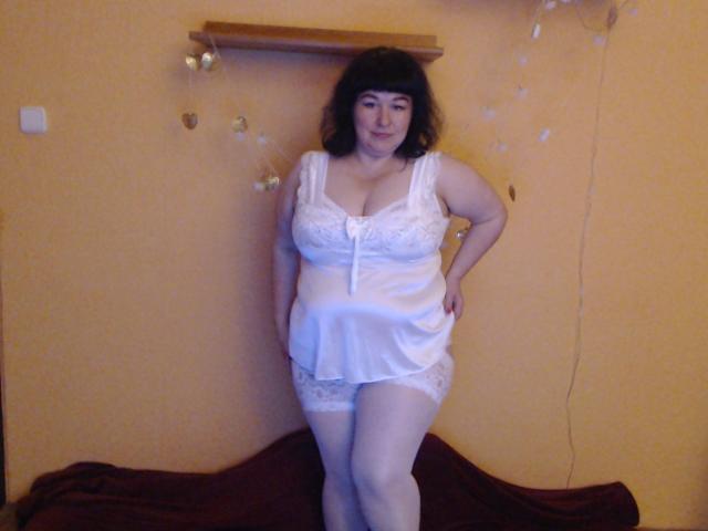 ErinSmart with curly black hair, white lace lingerie, and thigh-high stockings, standing with hands on hips, showing her big boobs and curvy ass. She's smiling slightly.