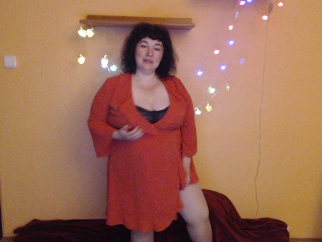ErinSmart, con cabello negro rizado, vestida con un vestido naranja, que deja ver lencería negra y sus senos, está de pie con una pierna levantada, hay un trozo de tela roja en el suelo y luces de colores..