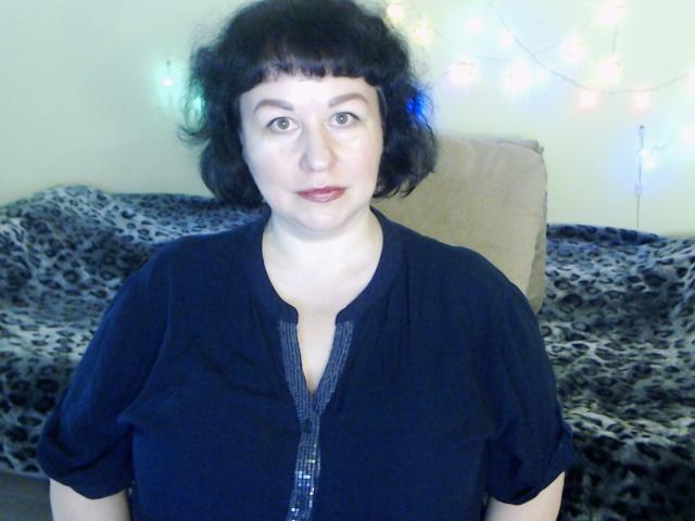 ErinSmart tem a pele clara, cabelo preto curto e encaracolado, e está vestindo uma camisa preta com botões na frente. Ela está olhando diretamente para a câmera com uma expressão neutra..