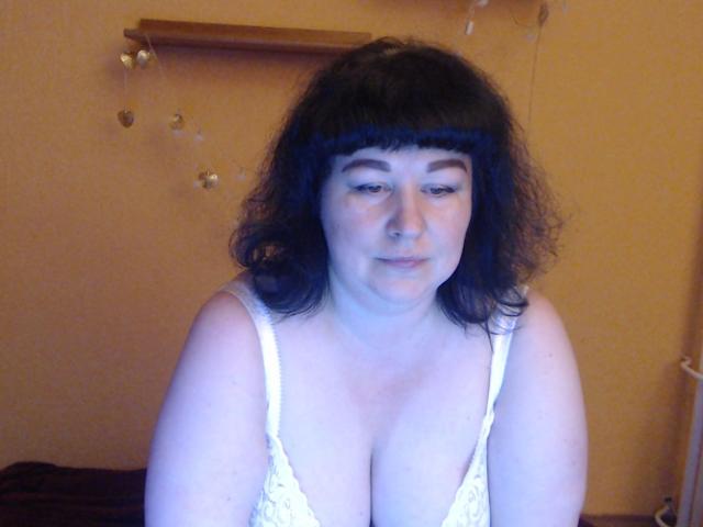 ErinSmart, con cabello negro rizado, un sostén de encaje blanco y senos grandes visibles, mirando hacia abajo con una ligera sonrisa, en una habitación con luz tenue..