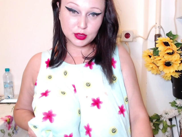 KissAndTits with dark hair, red lipstick, black eyeliner, wearing a light blue dress with pink flowers and green eyes, standing with large tits in a brightly lit room.