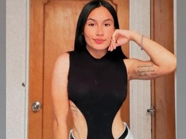 AshlyFerria with long black hair, wearing a black cut-out leotard, showing her side tattoos, arm raised, smiling, standing in front of a wooden door.
