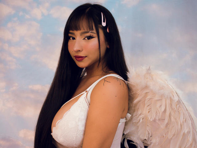 MiaSuarez with long black hair, white lace bra, and feather wings, looking over her shoulder, showing her tits, with a pink hairclip and red lipstick.