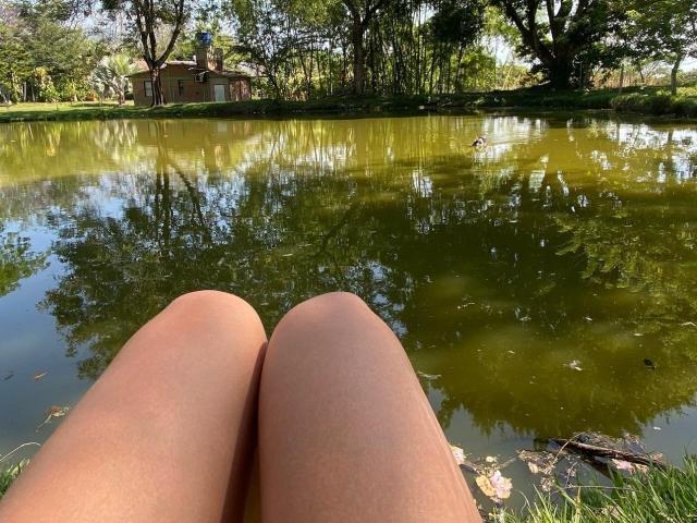 JuliaGarciax's legs are in the foreground, knees together, with a green-tinted, reflective pond in the background, showing tree reflections and a small red house.