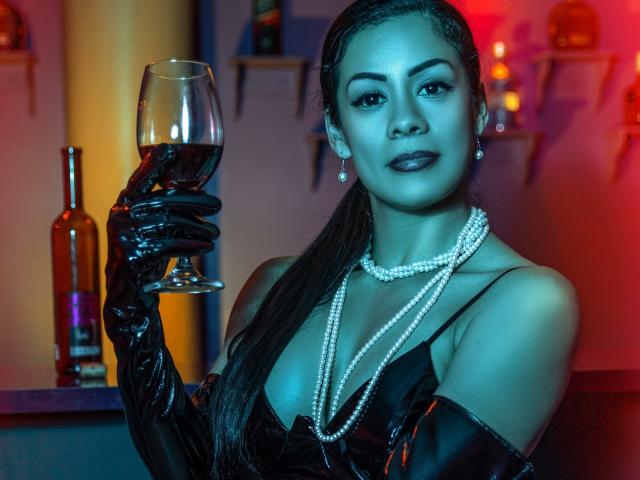 Nikkita Hernandez, con guanti di lattice neri, tiene in mano un bicchiere di vino rosso, indossa perle, rossetto scuro e ha i capelli lunghi, mostrando il seno in un bar sexy illuminato di blu e rosso..