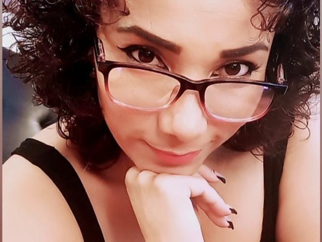 AmberDemons with curly black hair, wearing glasses and a black top, resting her chin on her hand, showing her dark nails and a subtle smile.