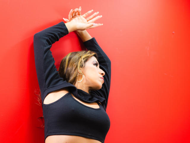 LaurenHills with arms raised, wearing a black crop top with cutouts, showing her tits, against a bright red wall, with gold hoop earrings, and side-swept blonde hair.