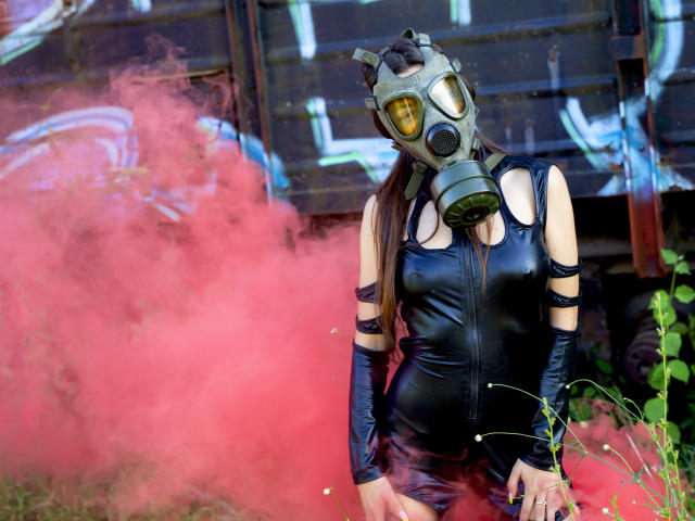AshlyChloe indossa un abito di lattice nero, una maschera antigas e guanti neri, ed è in piedi di fronte a del fumo rosso, con i suoi seni visibili attraverso dei tagli, all'aperto in una zona erbosa..