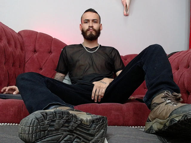 AndersX con la barba, una camicia nera trasparente, jeans neri e stivali marroni, gambe divaricate, seduto su un divano rosso, con tatuaggi visibili sulle braccia, che guarda la fotocamera, con un'espressione seria..