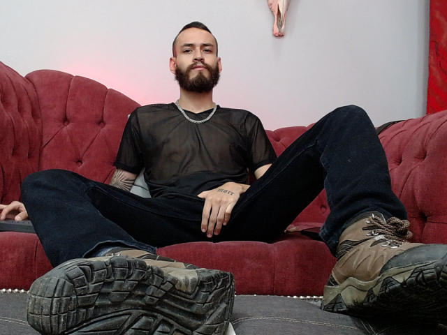AndersX, um homem de barba com uma tatuagem no braço, vestindo uma camisa preta transparente, calças escuras e botas sujas, sentado com as pernas abertas em um sofá vermelho, com uma expressão confiante..