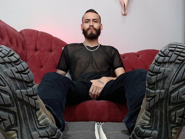 AndersX con barba, camisa negra transparente, pantalones negros, sentado en un sofá rojo, con las piernas separadas, mostrando botas sucias, tatuaje en la mano derecha, zapatillas blancas en el suelo..