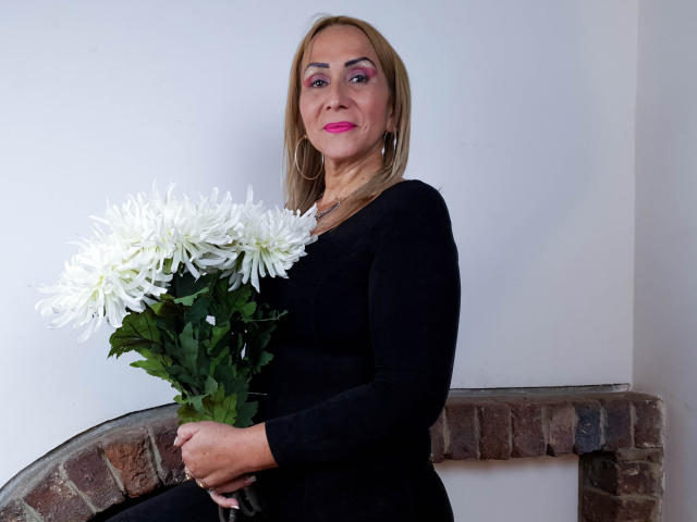 DianeOasis con lápiz labial rosa, sosteniendo crisantemos blancos, vestida con un vestido negro de manga larga, sentada en un banco de ladrillo, sonriendo, cabello rubio, sombra de ojos rosa, aros grandes..