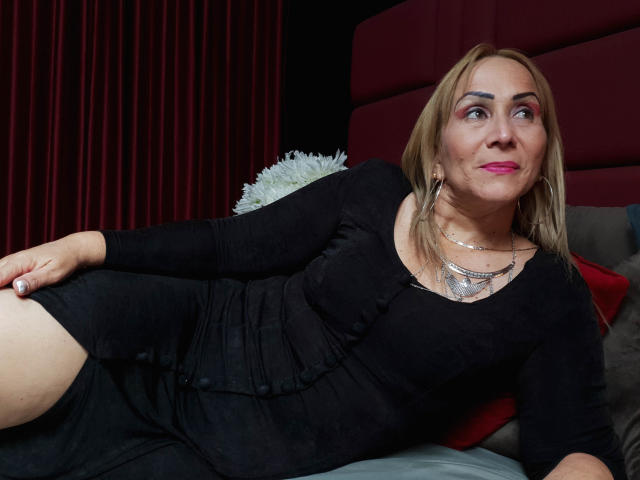 DianeOasis is a blonde woman with pink lipstick, wearing a black dress, lying on a bed with red and gray pillows, looking to the side, her hand on her thigh.