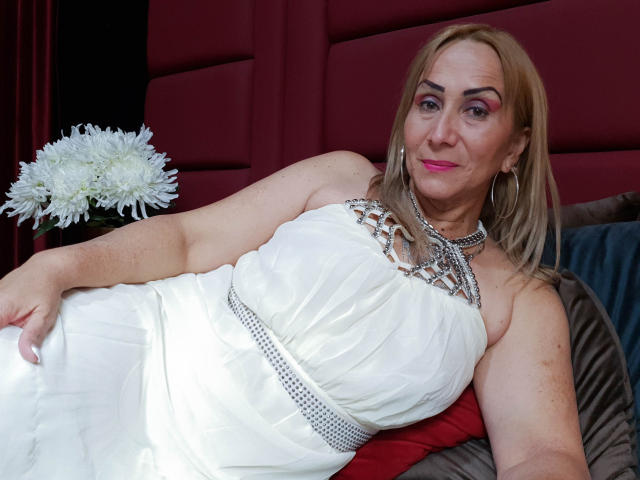 DianeOasis with blonde hair, pink lipstick, and hoop earrings, wearing a white dress with silver embellishments and a jeweled belt, laying on a red and gray cushion.