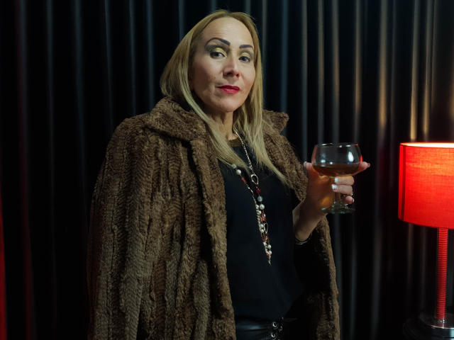 DianeOasis with blonde hair, wearing a brown furry coat, black top, and necklace, holding a glass of whiskey, red lamp on right, standing, confident, slightly smiling.