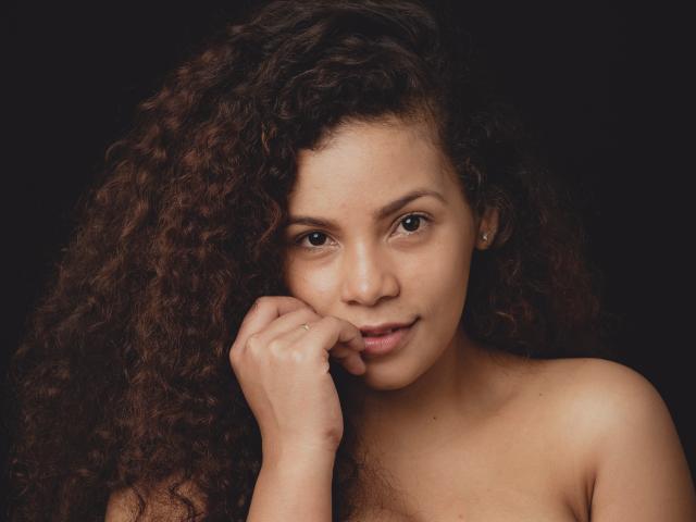 EbonyRose69 has curly brown hair, a light brown skin tone, and a soft smile with her finger gently touching her chin. She's topless with small tits and dark background.