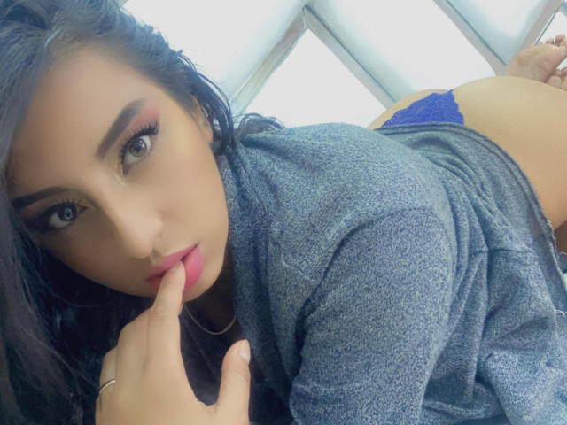 EmilyHarper with dark hair, pink eyelids, and gray top, biting finger, showing blue panties, and her ass, in a provocative pose, looking at camera.