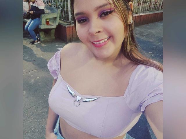 EvaMadison with small tits, smiling, wearing a pink off-shoulder top with a winged bow, purple eyeshadow, and blue jeans, standing on a street, taking a selfie.