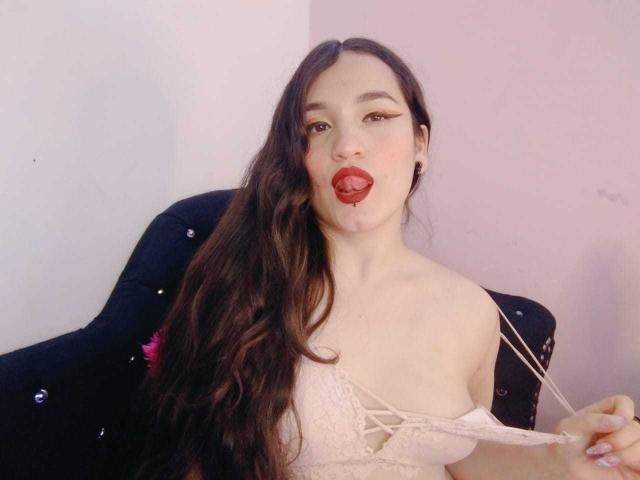 MillaDrew met lang bruin haar, tong uit, rode lipstick, witte kanten bh, die een bandje vast houdt, zittend op een zwarte stoel met glitters. Borsten zichtbaar. Bleke huid, subtiele make-up..