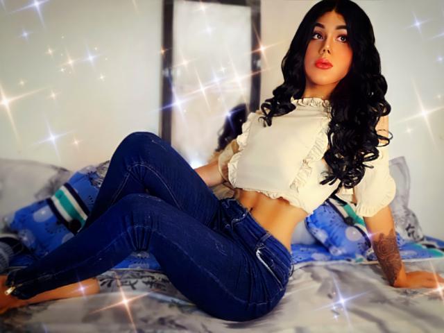 Isabella Veliz, met lang zwart haar, draagt een witte top met ruches en blauwe jeans, en zit op een bed met glitters. Ze laat haar getatoeëerde arm en rode lippen zien..