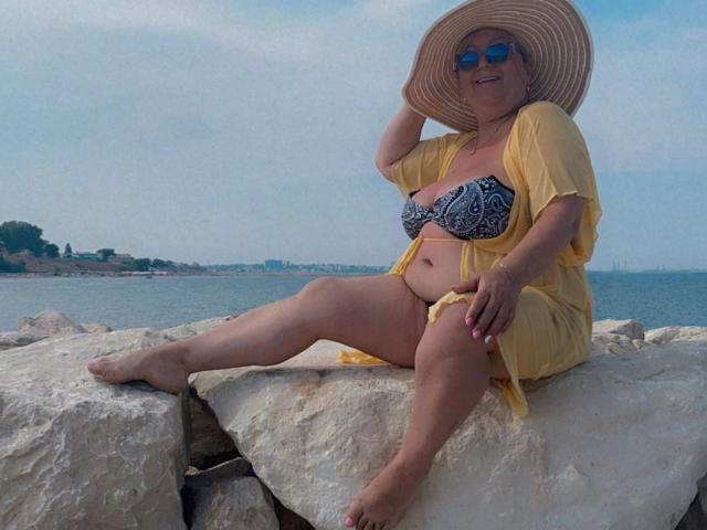 QueenPammy, chubby with big tits, in a black and white patterned bra, yellow open robe, and sun hat, sitting on rocks by the sea, wearing blue sunglasses.