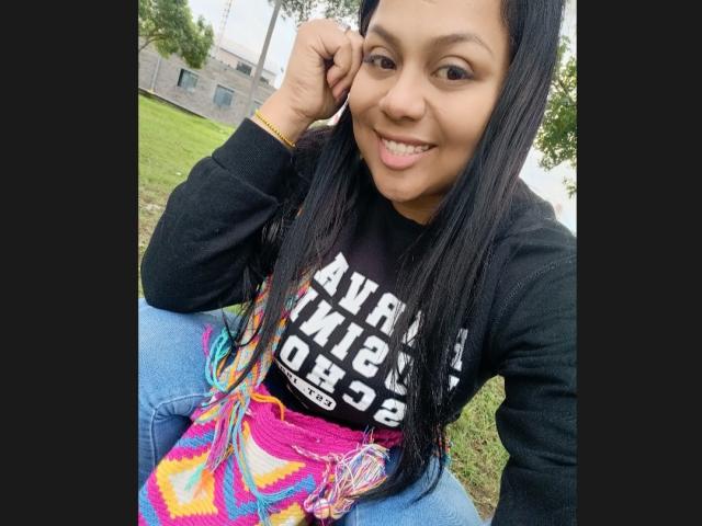 AvaSexySeins, avec de longs cheveux noirs, souriante, portant une chemise noire et une jupe colorée tie-dye, assise sur l'herbe, la main droite touchant son oreille, jean bleu, décor extérieur..