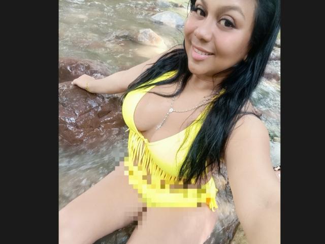 AvaSexySeins con cabello negro largo, parte superior de bikini amarilla con flecos y parte inferior de bikini amarilla censurada, sentada sobre rocas junto a un arroyo, sonriendo a la cámara..