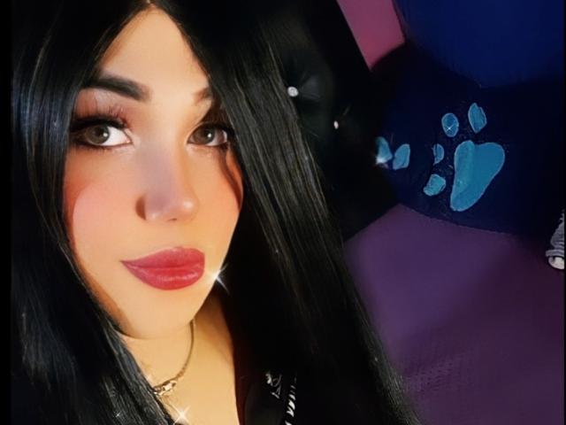 Isabella Veliz, com cabelos pretos longos, batom vermelho e olhos castanhos, vestindo uma blusa preta, tem um leve brilho na bochecha e um colar, olhando para a câmera..
