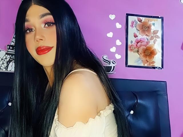 Isabella Veliz heeft lang zwart haar, rode lippenstift, roze oogschaduw, een witte top met een open schouder, waardoor haar blote schouder te zien is, en staat voor een paarse muur met een bloemmotief..