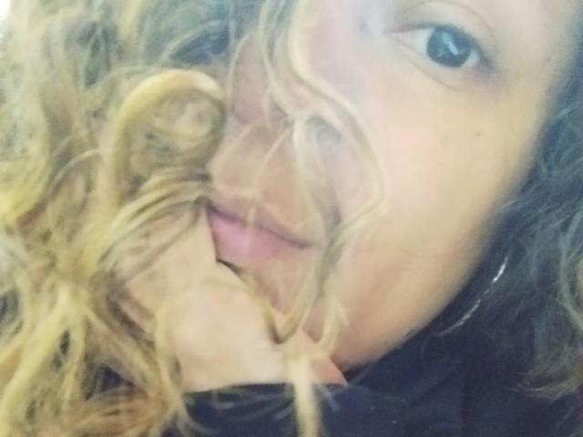 Angelonix's face is partially covered by her curly blonde hair, showing a slightly open mouth with a visible pink tongue. She's holding her hair with one hand.