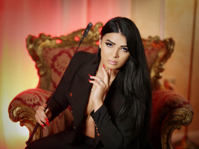 Devasha with long black hair, wearing a black suit, holding a whip, sitting on an ornate red chair, red nails, gold necklace, looking seductively at the camera.