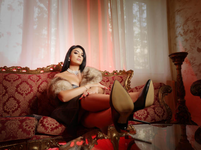 Devasha with black hair, wearing a fur stole, pearl necklace, and black high heels, sitting on a red and gold ornate couch, legs up, showing her tits and ass.