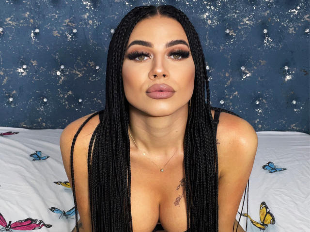 BriannaArons with long black braids, full pouty lips, and cleavage, wearing a black top, has a tattoo on her right chest, and a small necklace.