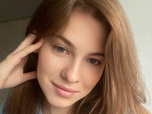 Das Gesicht von MineVicy, mit hellbraunen Haaren, blauen Augen und einem leichten Lächeln, zeigt eine Hand, die ihr Kopf berührt. Sie hat helle Haut und rosafarbene Lippen..
