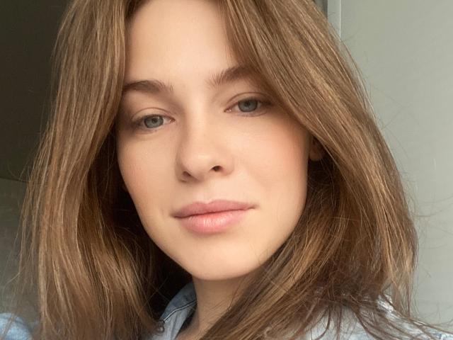 MineVicy ha i capelli castano chiaro, lisci, gli occhi verdi e le labbra rosate, con una carnagione chiara, indossa una camicia azzurra, in un selfie con una luce soffusa..