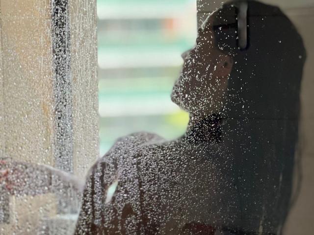 ValleryHottie com gotas de água em uma janela, virada para a esquerda, usando fones de ouvido, de costas para a câmera, cabelo escuro, ligeiramente borrada, molhada pela chuva, sentada dentro de casa, olhando para fora..