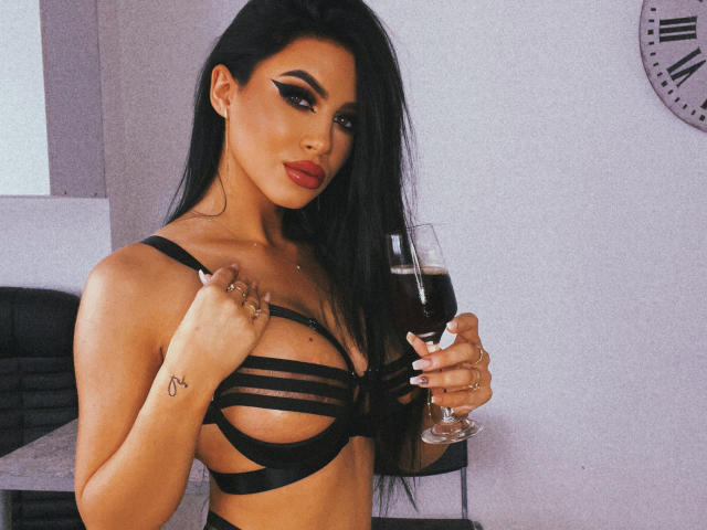 Brianna Arons mit langen, schwarzen Haaren, die schwarze, trägerlose Unterwäsche trägt, hält ein Glas Rotwein, zeigt ihre Brüste und hat ein Tattoo auf ihrer rechten Hand..