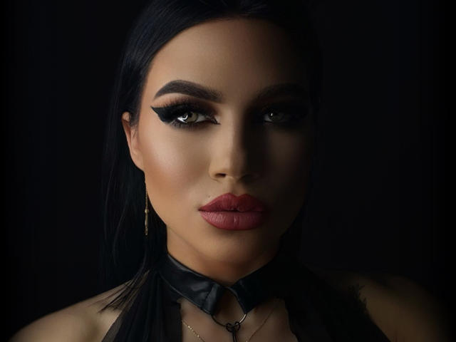 Brianna Arons tiene el cabello oscuro, cejas negras y gruesas, y lápiz labial rojo. Lleva delineador de ojos negro, un collar de cuero negro y aretes de aro dorados. Sus ojos son marrones y está mirando..